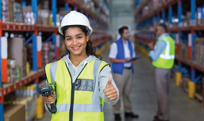 A woman wearing a safety vest and hard hat gives a thumbs up in a warehouse.