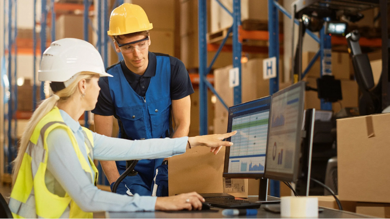 Two warehouse workers in safety gear discuss data displayed on a computer screen.