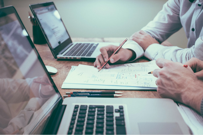 Two people collaborating on paperwork at a desk with laptops.