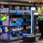 Two workers in a warehouse use autonomous robots to assist in organizing and managing inventory.