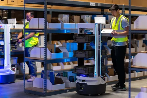 Two workers in a warehouse use autonomous robots to assist in organizing and managing inventory.