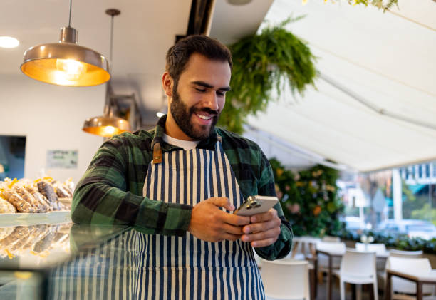 A smiling man in an apron checks his phone in a modern cafe, highlighting relaxed entrepreneurial spirit.