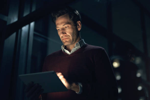 A business owner reviews potential fines on his tablet in a dimly lit office setting.