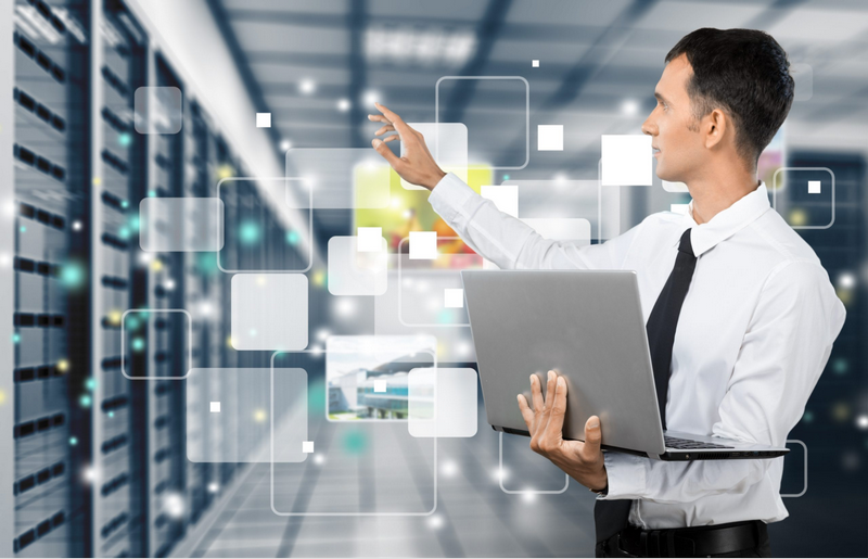 A man in a server room interacts with floating digital interfaces while holding a laptop.