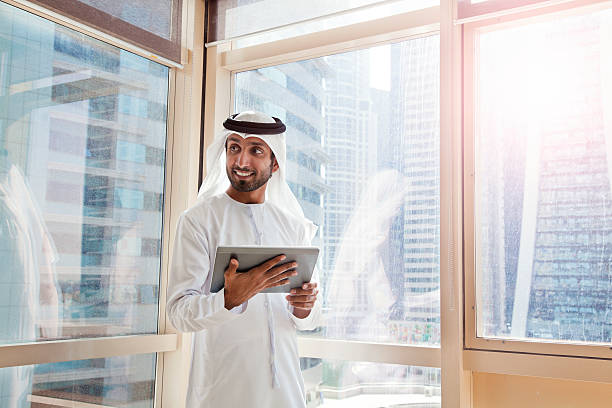 Businessman in traditional attire holds a tablet, standing near windows with city skyscrapers in the background.